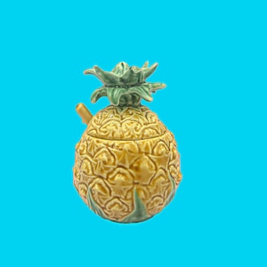 May include: A ceramic pineapple-shaped container with a lid and a small spoon. The container is primarily yellow with a textured surface, and the top has green leaves. The background is a solid blue.