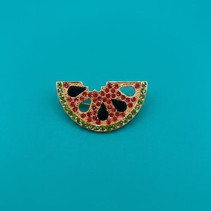 May include: A gold-tone brooch shaped like a watermelon slice, featuring a row of green crystals along the curved edge, a center section filled with small red crystals, and three black teardrop-shaped accents representing seeds.