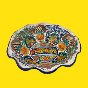 May include: A decorative ceramic bowl with a scalloped edge, featuring a vibrant hand-painted floral design. The bowl's interior showcases a central oval with intricate patterns in blue, orange, and green. The rim is a deep blue, and the background is yellow.