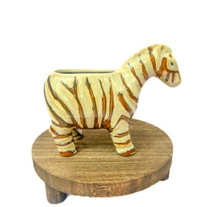 May include: A ceramic zebra planter with brown and white stripes. The planter is sitting on a wooden stand with three legs.