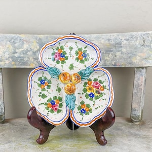 May include: A white ceramic serving dish with three sections, each decorated with floral and fruit motifs in shades of blue, yellow, orange, and green. The dish is shaped like a clover leaf and has a scalloped edge.