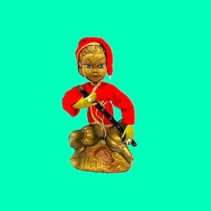May include: A whimsical figurine of a seated figure in a red outfit and hat, holding a black instrument. The figure has a light-colored face and hands, with gold accents and a gold base. The background is a solid teal color.