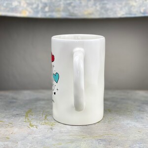 Pu&ograve; includere: Tazza in ceramica bianca con un design a forma di cuore blu e rosso e un manico bianco. La tazza ha una grafica in bianco e nero sul davanti.