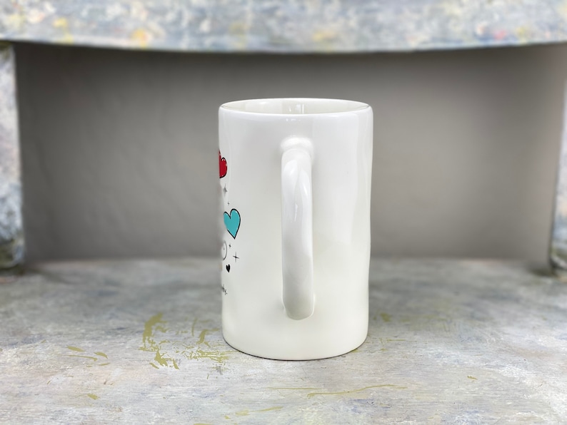 Pu&ograve; includere: Tazza in ceramica bianca con un design a forma di cuore blu e rosso e un manico bianco. La tazza ha una grafica in bianco e nero sul davanti.