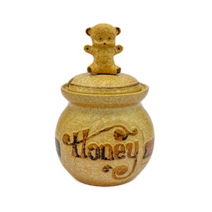 May include: A ceramic honey pot with a bear-shaped lid. The pot is decorated with the word "Honey" in a brown script.