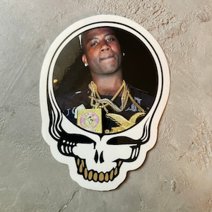 Puede incluir: Una pegatina en blanco y negro de una calavera con una cara sonriente. La calavera tiene un contorno negro y dientes dorados. Dentro de la calavera hay una foto de un hombre con un collar de cadena de oro.