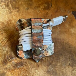 May include: A white cord wrapped around a leather cord organiser with a snakeskin pattern. The organiser has a metal button closure and is in a light brown wooden bowl.