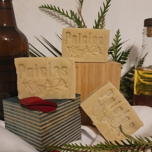 Puede incluir: Tres barras de jabón natural con la palabra "Paiclas" en relieve en cada barra. Las barras están apiladas una encima de la otra sobre una superficie de madera.