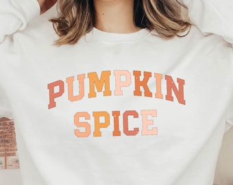 May include: A white sweatshirt with the text "Pumpkin Spice" in a colorful, layered font. The sweatshirt is worn by a person with long brown hair and a brown hat.