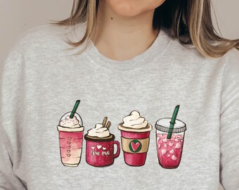 Puede incluir: Una sudadera gris con un gráfico de cinco tazas de café y bebidas heladas diferentes. Las tazas son todas rosas y blancas con detalles verdes. El texto "Love Me" está en una de las tazas.