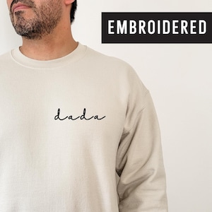 Könnte beinhalten: Ein beiger Pullover mit dem Wort "dada" in schwarzer kursivschrift auf der Vorderseite gestickt. Der Pullover ist ein Rundhalsmodell mit langen Ärmeln.