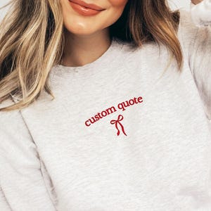 Peut inclure: Un sweat-shirt gris clair avec l'inscription "custom quote" en rouge, accompagné d'un motif de nœud rouge. Le sweat-shirt a un col rond et des manches longues.