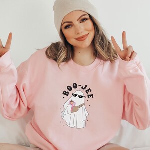 May include: A pink sweatshirt with a ghost wearing sunglasses and holding a drink. The ghost is surrounded by stars and the text "BOO-JEE" is written in black letters.