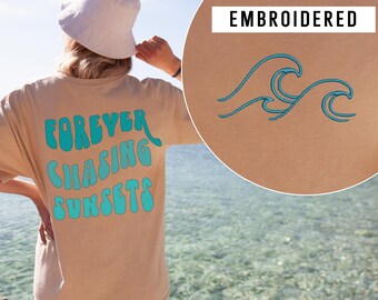 May include: Tan t-shirt with the text "FOREVER CHASING SUNSETS" in turquoise. The shirt has a relaxed fit. A close-up shows an embroidered turquoise wave design. The shirt is a casual, beach-themed apparel item.