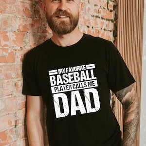 May include: Black t-shirt with the text "MY FAVORITE BASEBALL PLAYER CALLS ME DAD" in white, distressed font. The shirt is worn by a person with a beard, leaning against a brick wall. The overall theme is Father's Day or baseball.