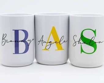 Könnte beinhalten: Drei weiße Keramikbecher mit personalisierten Namen und Anfangsbuchstaben. Die Becher sind in einer Reihe von links nach rechts angeordnet. Der erste Becher hat den Namen "Bronwyn" mit einem violetten "B". Der zweite Becher hat den Namen "Angela" mit einem gelben "A". Der dritte Becher hat den Namen "Shalon" mit einem grünen "S".