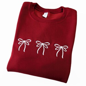 Könnte beinhalten: Ein gefaltetes weinrotes Sweatshirt mit drei weißen Schleifenmotiven, die auf der Vorderseite gestickt sind. Das Sweatshirt hat einen Rundhalsausschnitt und ein kleines Etikett im Kragen.