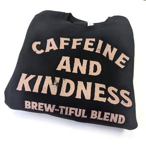 May include: Black sweatshirt with the words "CAFFEINE AND KINDNESS" in a light brown, distressed font. Below the words is the phrase "BREW-TIFUL BLEND". The sweatshirt is folded on a white surface.