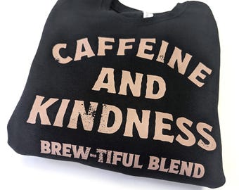 May include: Black sweatshirt with the words "CAFFEINE AND KINDNESS" in a light brown, distressed font. Below the words is the phrase "BREW-TIFUL BLEND". The sweatshirt is folded on a white surface.