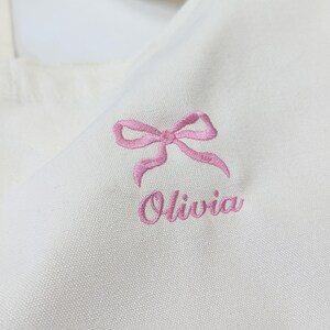 May include: A close-up of a white tote bag featuring a pink embroidered bow and the name "Olivia" in a matching pink script. The bag is made of a textured fabric.