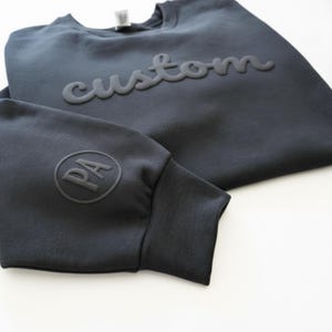 May include: A navy blue sweatshirt with the word "custom" in raised lettering across the chest. The left sleeve features a circular emblem with the letters "PA". The sweatshirt is made of a soft material.