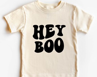 Può includere: Una maglietta color crema a maniche corte con il testo "HEY BOO" in lettere retrò nere.