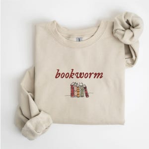 May include: A beige sweatshirt with the text "bookworm" in red cursive lettering and a small embroidered image of a stack of colorful books.