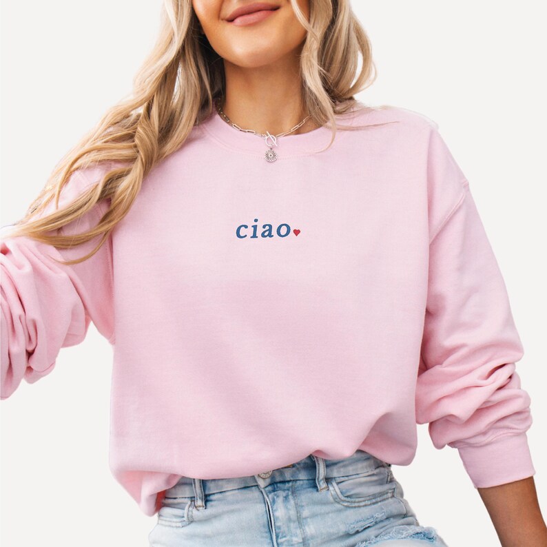 May include: A light pink sweatshirt with the word "ciao" in blue, with a red heart. The sweatshirt has long sleeves and a crew neck. The garment is made of a soft material.