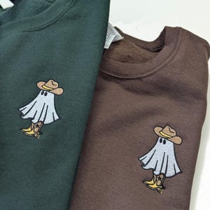 Könnte beinhalten: Zwei Sweatshirts, eines dunkelgrün und eines braun, mit einem gestickten Design eines Geistes, der einen Cowboyhut und Stiefel trägt.