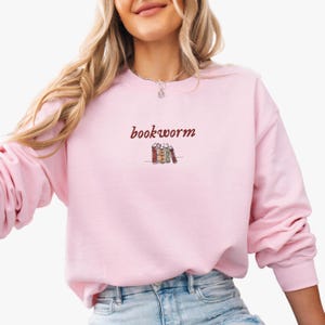 May include: A pink sweatshirt with the text "bookworm" in maroon lettering and a small illustration of a stack of books.