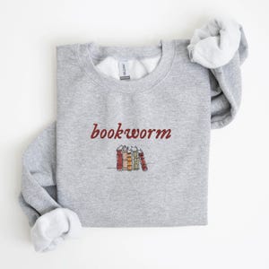May include: Gray sweatshirt with the text "bookworm" in red cursive lettering and a small illustration of a stack of books below the text.