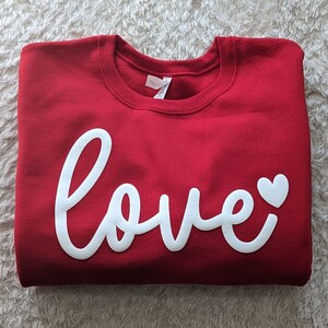 May include: Red sweatshirt with a white cursive "love" with a small white heart.