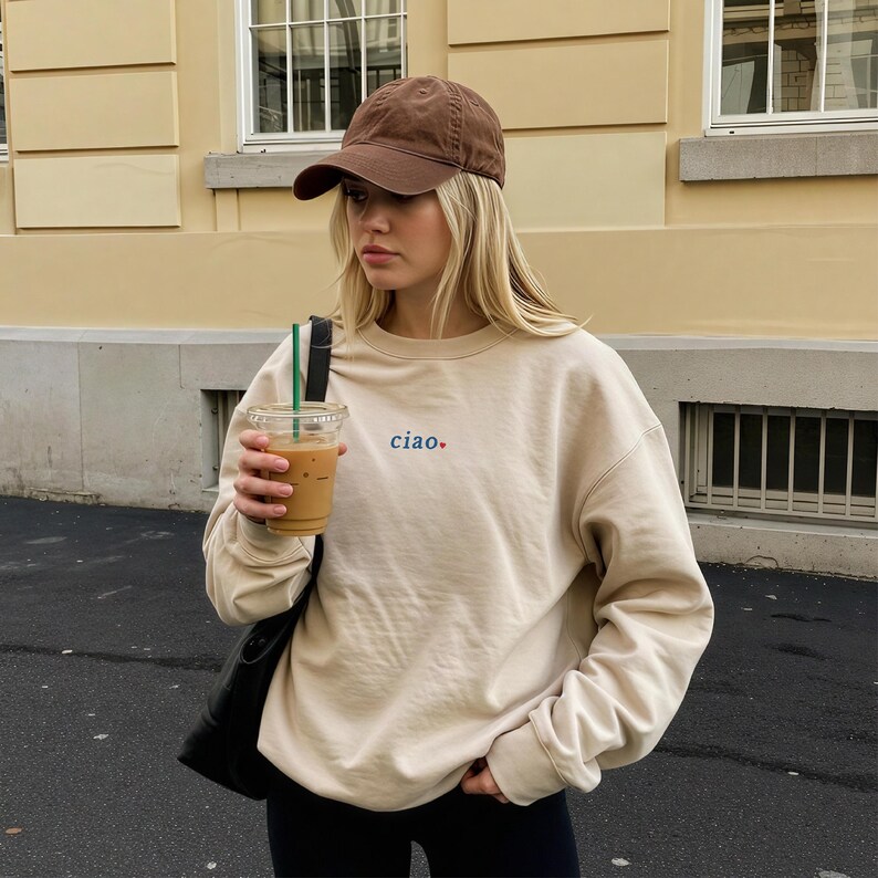 May include: A beige sweatshirt with the word "ciao" embroidered in blue and red. The person in the image is wearing a brown baseball cap and holding a clear plastic cup with a beverage and a green straw. A black shoulder bag is also visible.
