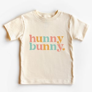 May include: A cream colored short-sleeved t-shirt with the text "hunny bunny" in colorful letters. The letters are in shades of pink, orange, and blue.