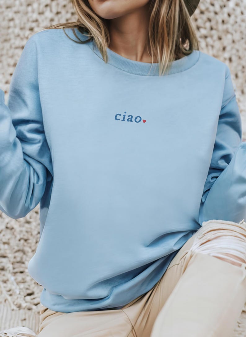 May include: A light blue sweatshirt with long sleeves. The word "ciao" is embroidered in blue with a small red heart. The sweatshirt is made of a soft material and is a casual style.
