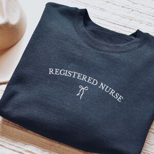 Könnte beinhalten: Ein marineblaues Sweatshirt mit dem weißen Schriftzug "REGISTERED NURSE", der sich bogenförmig über der Brust erstreckt, und einer kleinen Schleife. Ein cremefarbener Hut und Schmuck sind im Hintergrund zu sehen.