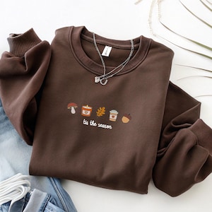 Peut inclure: Un sweat-shirt marron avec un col rond et des manches longues. Le devant présente un motif brodé d'un champignon, d'une bougie, d'une feuille, d'une tasse à café et d'un gland. Le texte "tis the season" est en dessous. Un collier argenté est posé dessus.