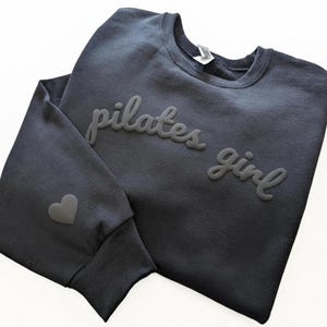 Op de afbeelding: Een marineblauw sweatshirt met de woorden "pilates girl" in een verhoogd, gestructureerd lettertype. Er staat een klein hartje op de linkermouw. Het sweatshirt is opgevouwen en toont de voorkant en een deel van de mouw.