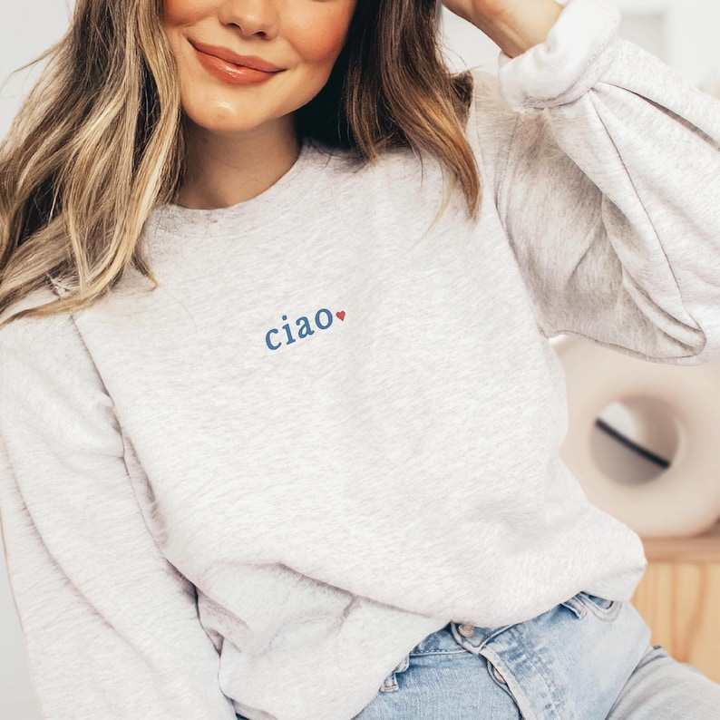 May include: A light gray sweatshirt with the word "ciao" embroidered in blue, with a red heart. The sweatshirt has long sleeves and a crew neck. The model is wearing blue jeans.