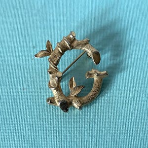 May include: A gold-toned metal brooch shaped like the letter G, designed with a textured, bark-like finish that mimics the appearance of a tree branch. The letter features small, pointed leaves attached to the stem and a pin mechanism on the back.