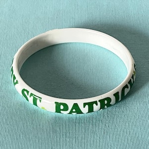 Könnte beinhalten: Ein weißer Armreif mit grünem Schriftzug ST. PATRICK'S DAY und kleinen grünen Kleeblatt-Symbolen, die auf die Außenseite des kreisförmigen Bandes gedruckt sind. Der Armreif hat eine glatte, glänzende Oberfläche und ein schlichtes Design.
