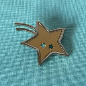 May include: A gold-colored star-shaped pin with a silver border and two small teal stars inside. The pin has two curved silver lines extending from the top, suggesting motion. The pin is set against a teal background.