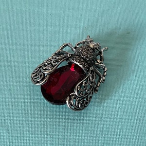 May include: A silver-toned bee brooch with intricate filigree wings and a large, oval, ruby-red gemstone body. The bee's head is detailed with small, dark-colored stones. The brooch is set against a light blue background.