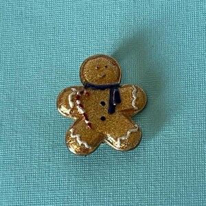 May include: A gold gingerbread man pin with white icing and a red and white candy cane. The gingerbread man is wearing a blue scarf.