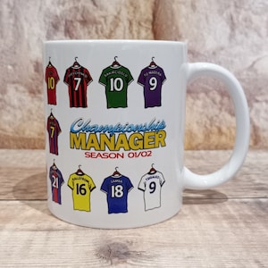 May include: White ceramic mug featuring a design of football shirts with numbers and names. The text "Championship Manager Season 01/02" is printed on the mug.