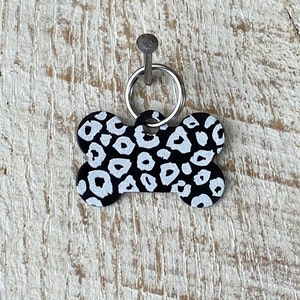 May include: Black and white bone-shaped dog tag with a leopard print design. The tag has a silver ring for attaching to a collar.