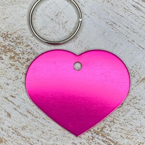 May include: A bright pink heart-shaped metal tag with a hole for a key ring. The tag is on a light wood background.