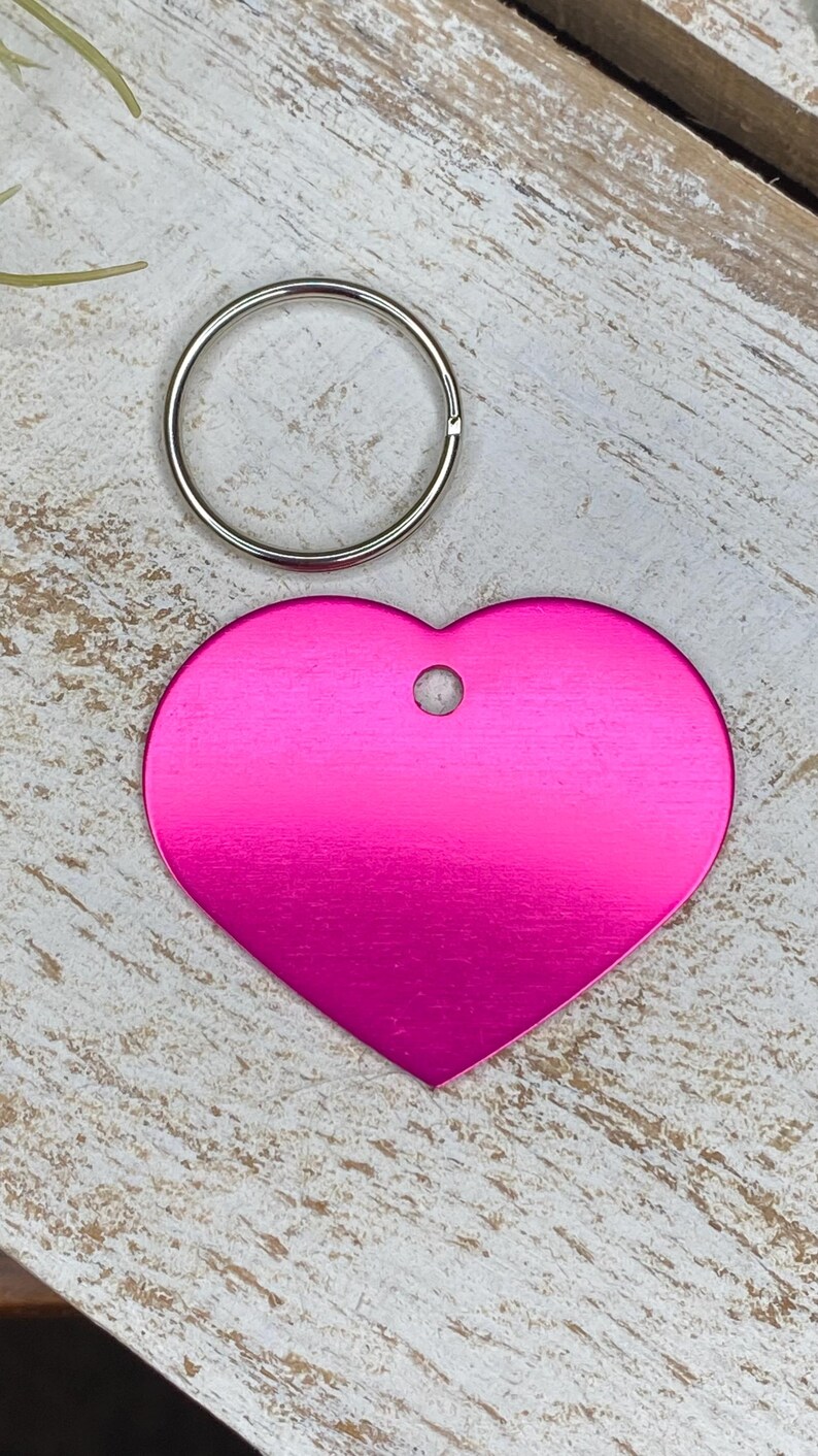 May include: A bright pink heart-shaped metal tag with a hole for a key ring. The tag is on a light wood background.