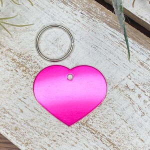 May include: A pink heart-shaped metal tag with a hole for a ring. The tag is on a light wood surface.