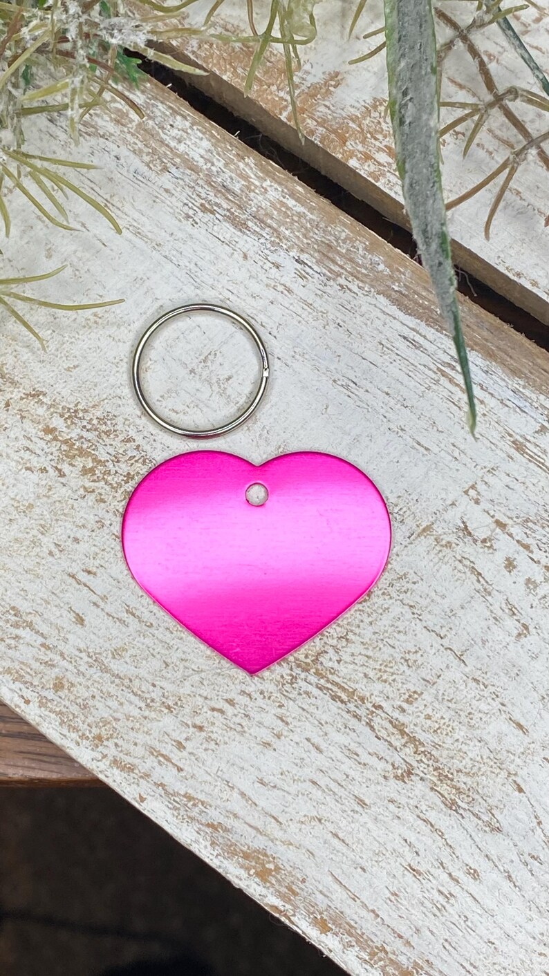 May include: A pink heart-shaped metal tag with a hole for a ring. The tag is on a light wood surface.